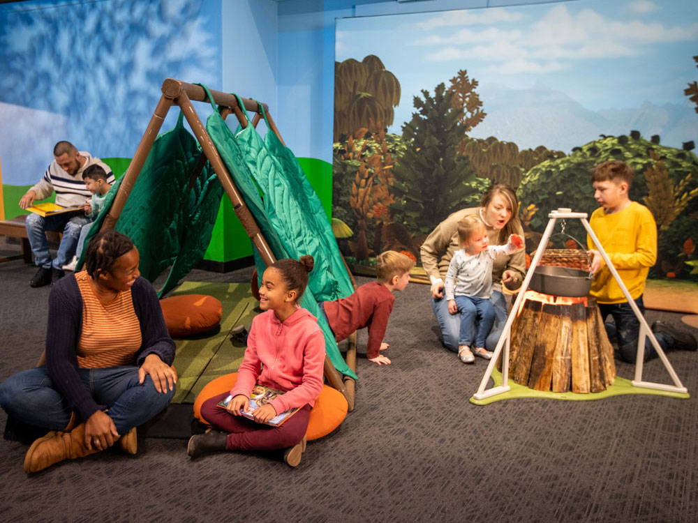 An adult and child are sitting in front of a green tent. A child is crawling out of the tent. Another family is looking at an object that looks like a tripod built over a campfire.